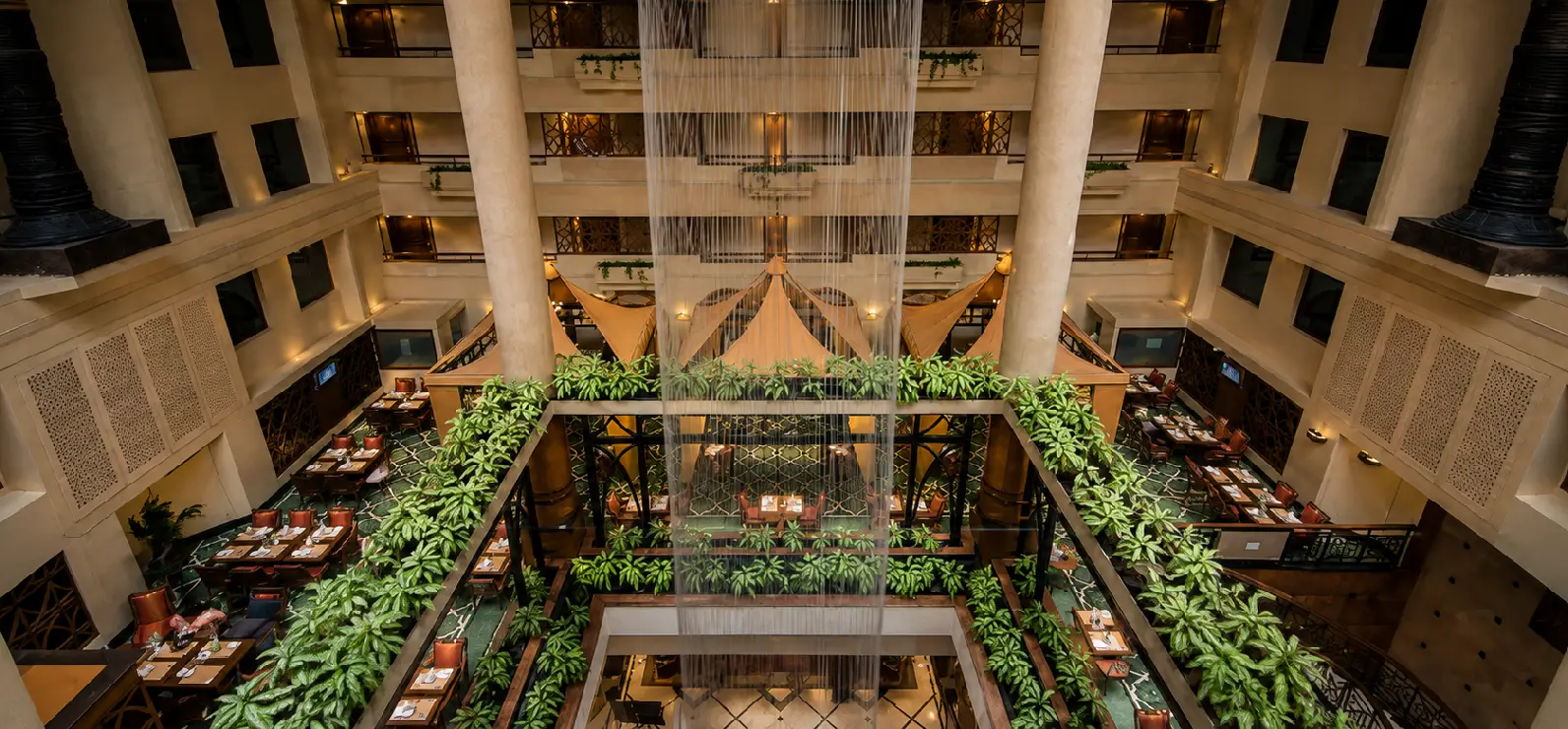 The Orchid Hotel Mumbai Atrium - Festive Staycation Venue Grand multi-tier atrium balcony corridor featuring ornate wrought-iron railings, lush indoor atrium plants, and an indoor fountain at The Orchid Hotel Vile Parle